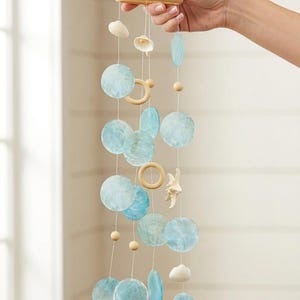 Puede incluir: Móvil colgante decorativo con elementos redondos y ovalados de color azul claro, pequeñas cuentas de madera, anillos de madera y conchas marinas. El móvil está suspendido de un anillo de madera natural. Tema costero.
