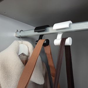 Op de afbeelding: Een zwarte en een witte S-vormige kledinghaak hangen aan een metalen stang. De zwarte haak ondersteunt een beige riem, terwijl de witte haak een donkerbruine riem ondersteunt. Een beige jas met een witte imitatiebontkraag hangt aan een witte hanger.