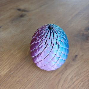Könnte beinhalten: Ein ovales Objekt mit einer strukturierten, schuppenartigen Oberfläche. Der Gegenstand weist einen Farbverlauf auf, der von einem tiefen Magenta- und Violettton auf der einen Seite zu einem leuchtenden Cyan- und Blauton auf der anderen Seite übergeht.