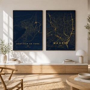 Puede incluir: Dos impresiones enmarcadas de mapas de ciudades. Una presenta Santiago de Cuba, Cuba, y la otra Madrid, España. Los mapas son dorados sobre un fondo azul oscuro. Las impresiones se exhiben en una pared blanca sobre un mueble de madera.