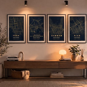 Op de afbeelding: Vier ingelijste kunstprints van stadskaarten hangen boven een houten console. De kaarten, in goud op een donkerblauwe achtergrond, tonen de steden Yeda, Makkah Al-Mukarramah, Dammam en Riad, Saoedi-Arabië. Een lamp, boeken en planten sieren de tafel.