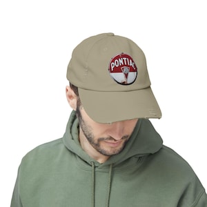 May include: A beige baseball cap with a distressed Pontiac logo. The logo is circular, with a red and white background and the word "PONTIAC" arched above. The cap has a worn, vintage appearance, with a slightly frayed peak.