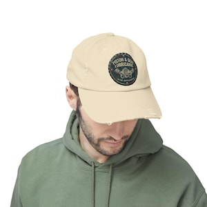 May include: A cream-coloured distressed baseball cap. It features a circular emblem with the text "Piston & Gear Lubricants" and an engine parts graphic. The cap has a worn appearance with frayed edges.
