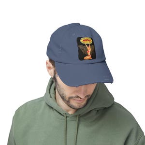 Puede incluir: Gorra de béisbol azul desgastada con un gráfico de estilo vintage. El gráfico incluye la palabra "Mopar" y una ilustración de estilo pin-up. La gorra tiene una visera curva y un aspecto desgastado.