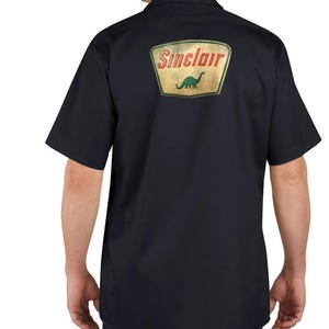 May include: Black short-sleeve work shirt with a Sinclair logo patch on the back. The patch features the Sinclair name in red and a green dinosaur graphic on a yellow and green background.
