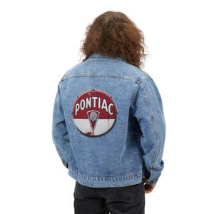 May include: Light blue denim jacket with a large, circular Pontiac logo on the back. The logo features the word "Pontiac" in white, a red background, and a Native American chief graphic.