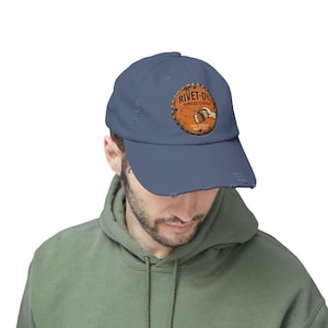 May include: A distressed blue baseball cap featuring a circular patch with the text "RIVET-OY SERVICE STATION" and an illustration of a hand holding a can. The cap has a pre-worn look.