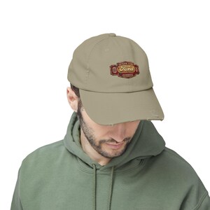 May include: A tan baseball cap with a distressed brim. The cap features a Ford logo in red and gold with the text "GENUINE FORD" and "AUTHORIZED DISTRIBUTOR". The cap is worn by a person wearing a green hoodie.