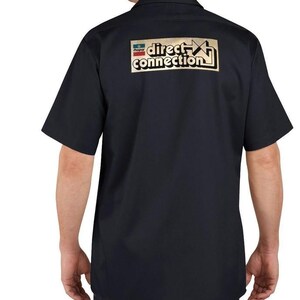 Pode incluir: Camisa de trabalho preta de manga curta com o logotipo "direct connection" dourado e vermelho nas costas. A camisa é de cor preta lisa e tem gola abotoada.