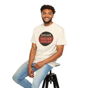 May include: Cream-colored t-shirt featuring a Sinclair Opaline Motor Oil logo in a black, red, and gold oval design. The shirt is worn by a person sitting on a black stool, wearing blue jeans.
