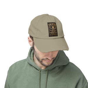 Puede incluir: Gorra de béisbol color beige con visera desgastada. La gorra presenta un parche rectangular con la palabra "MOPA" sobre un emblema de estrella y las palabras "Quality Performance" y "Just Drive It And Feel The Difference."