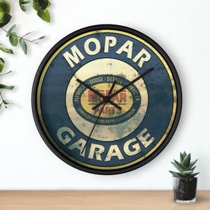 May include: Round wall clock with a black frame featuring the MOPAR Garage logo in white and yellow on a blue background. The clock face includes the names of car brands like Dodge and Chrysler.