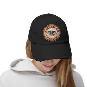 May include: Black distressed baseball cap with a circular emblem displaying "AMERICAN GASOLINE" and an eagle. The cap has a worn appearance with frayed edges, giving it a vintage look. The cap is worn by a person.