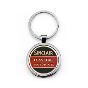 May include: A round, silver keychain with a circular pendant featuring the Sinclair Opaline Motor Oil logo. The logo is in red, black, and gold, with the text "SINCLAIR" at the top, "OPALINE" in the middle, and "MOTOR OIL" at the bottom.