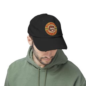 May include: Black distressed baseball cap with a circular Gilmore Gasoline logo featuring a lion graphic in red, yellow, and brown. The cap has a curved peak and a distressed finish.