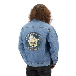 May include: A light blue denim jacket with a circular graphic on the back. The graphic features a cartoon pig wearing a hat, with the words "Piggley Wiggley The Original Self Service" in a circular design. The jacket has a classic design.