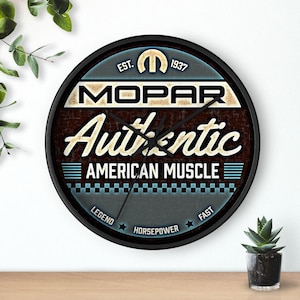 May include: Round wall clock with a black frame. The clock face features the text "MOPAR Authentic American Muscle" in white and cream against a brown and blue background. The clock also has the text "EST. 1937".