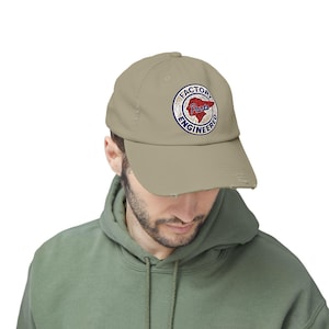 May include: A beige baseball cap with a worn-out peak. The cap has a circular emblem with the words "FACTORY ENGINEERED" around a red heart shape with "Paris" in the centre. The cap is worn by a person.