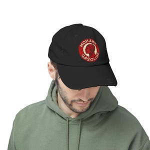 May include: Black distressed baseball cap with a red and white circular logo. The logo features the text "MOHAWK GASOLINE" and a Native American profile. The cap has a curved peak and a worn, vintage look.