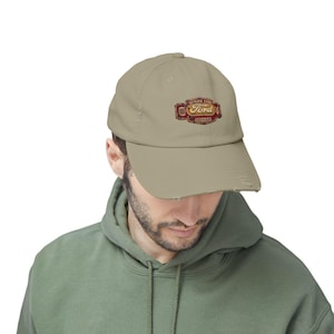 May include: A tan baseball cap with a distressed brim. The cap displays a Ford logo in red and gold, with the text "GENUINE FORD" and "AUTHORIZED DISTRIBUTOR". The cap is worn by a person wearing a green hoodie.