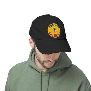May include: Black baseball cap with a distressed brim. The cap features a circular logo with the words "CONOCO GASOLINE" and an illustration of a man in a yellow circle.