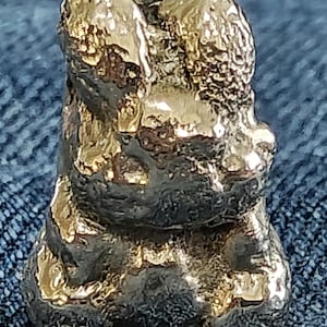 May include: A small, three-dimensional figurine shaped like a rabbit, crafted from a metallic material with a textured, uneven surface. The object features a reflective gold-toned finish with darker, recessed areas that highlight its organic form.
