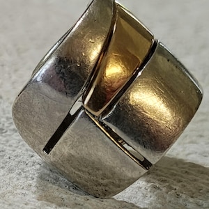 May include: A ring featuring a wide band constructed from alternating segments of polished silver-toned metal and smooth gold-toned metal. The design consists of interlocking rectangular panels that create a continuous, thick, and solid band structure.