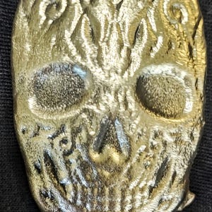 May include: A detailed, metallic skull pendant with intricate embossed patterns. The skull features prominent eye sockets, a defined nose, and a detailed jawline. The pendant has a textured, aged appearance.