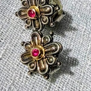 Puede incluir: Un par de pendientes de botón plateados con forma de flor. Cada pendiente presenta un centro dorado con una pequeña gema roja redonda. Los pétalos están detallados con pequeñas cuentas plateadas.