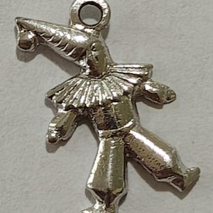 May include: A silver-toned metal charm shaped like a jester wearing a pointed hat with a small ball at the tip, a ruffled collar, and loose-fitting pants. The figure is depicted in a dynamic pose with one leg extended and includes a circular loop at the top.