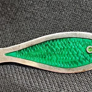 Op de afbeelding: Zilverkleurige visvormige broche met een getextureerde groene emaille body en een klein groen edelsteen oog. De broche heeft een gedetailleerd ontwerp, tegen een donkere achtergrond.