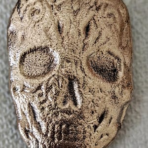 May include: A bronze-coloured skull pendant with detailed embossed patterns. The skull has visible eye sockets, a nasal cavity, and a detailed jawline. The surface has a textured look, and the design suggests a Day of the Dead theme.