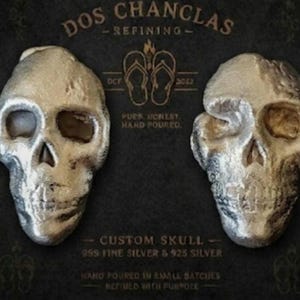 May include: Two silver skull sculptures with detailed facial features are displayed against a dark background. The text "DOS CHANCLAS - REFINING" and "CUSTOM SKULL" are visible. The skulls are made of .999 fine silver and .925 silver.