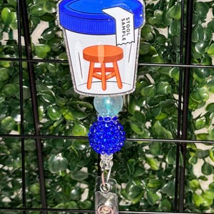 May include: A badge reel featuring a clear plastic container with a blue lid and a white label that reads STOOL SAMPLE. Inside the container is an orange stool illustration. The reel is decorated with a light blue bead and a textured dark blue bead.