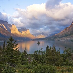 May include: A scenic landscape featuring a calm lake surrounded by mountains and evergreen trees. The mountains are illuminated by sunlight, with clouds overhead. The scene evokes a sense of tranquility and natural beauty.