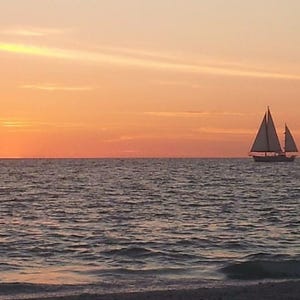May include: A sailboat with two white sails is silhouetted against a vibrant orange and yellow sunset over the ocean. The water is dark and rippled, with a sandy beach visible in the foreground.