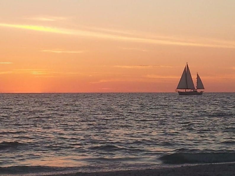 May include: A sailboat with two white sails is silhouetted against a vibrant orange and yellow sunset over the ocean. The water is dark and rippled, with a sandy beach visible in the foreground.