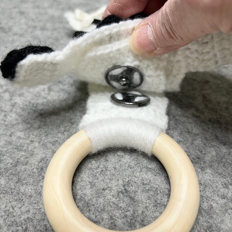 以下が含まれることがあります： 黒いアクセントが付いた白いかぎ針編みの布地で、銀色の金属製スナップボタンが付いています。布地は白い糸で巻かれた滑らかで明るい色の木製リングに取り付けられています。スナップボタンは開いた状態で、金属部分が見えています。