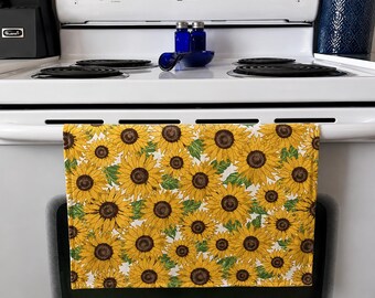 Op de afbeelding: Een keukendoek met een herhaald patroon van gele zonnebloemen met bruine centra en groene bladeren tegen een witte achtergrond. De handdoek hangt aan de ovendeur. Het thema is keukendecoratie.