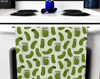Op de afbeelding: Een lichtgroene keukendoek met een herhaald patroon van cartoonaugurken met zonnebrillen en augurken in potten. De handdoek hangt aan een witte ovendeurknop.