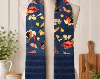 Op de afbeelding: Een hangende keukendoek met een donkerblauwe achtergrond en een herhalend patroon van hanen, hennen en kuikens in rood-, oranje- en geeltinten. Het onderste gedeelte bestaat uit een effen donkerblauwe, geribbelde badstof.