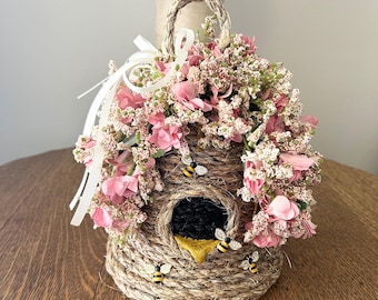 Op de afbeelding: Een decoratief bijenkorfvormig ornament gemaakt van geweven touw, versierd met roze en witte bloemen. De korf heeft een zwarte opening en is versierd met kleine bijenbeeldjes. Een geweven handvat en een wit lint maken het ontwerp compleet.