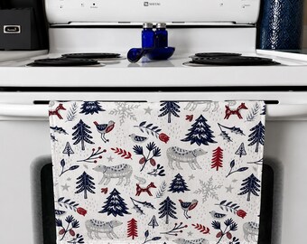Op de afbeelding: Witte keukendoek met een winterbospatroon. Het ontwerp omvat grijze beren, rode en blauwe bomen, vogels en sneeuwvlokken. De handdoek hangt aan een wit fornuis.
