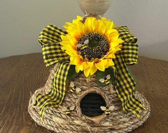 Op de afbeelding: Een decoratief vogelhuisje met een zonnebloem en een zwart-gele geruite strik. Het vogelhuisje is gemaakt van geweven touw en heeft een ronde opening. Decoratieve bijenaccenten zijn rond het vogelhuisje geplaatst.
