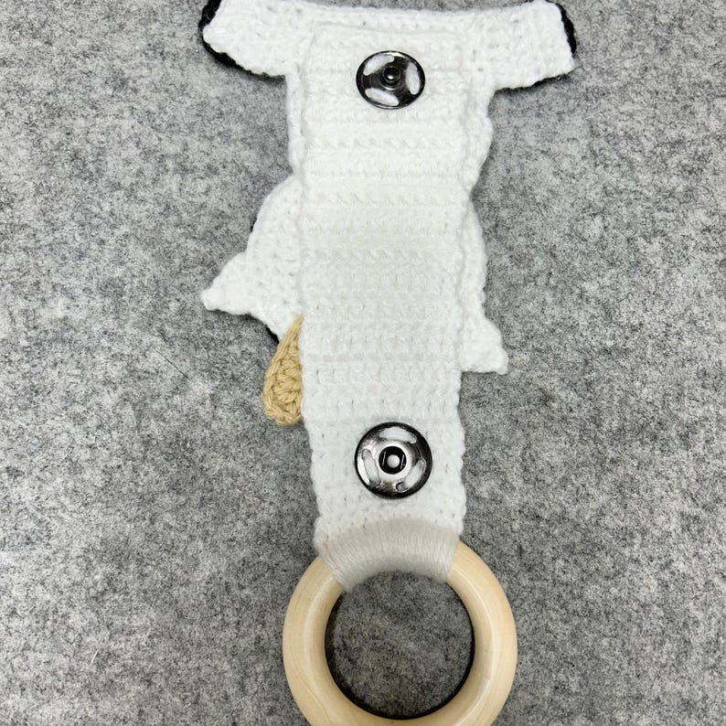 以下が含まれることがあります： 2つの銀色の金属製スナップボタンと側面に薄茶色のクロシェ編みのアクセントが付いた、白いクロシェ編みのストラップ。ストラップの下部には滑らかな円形の明るい色の木製リングが取り付けられており、機能的なホルダーになっています。