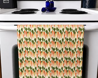 Op de afbeelding: Een keukendoek met een herhaald patroon van aquarelwortelen in tinten oranje, geel en groen. De wortelen zijn verticaal gerangschikt op een witte achtergrond. De handdoek hangt aan een witte oven.