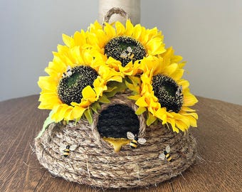 Op de afbeelding: Een decoratief bijenkorfvormig ornament gemaakt van geweven touw, versierd met felgele zonnebloemen en kleine bijenversieringen. De korf heeft een donkere opening en een gouden druppel. Een rustiek en charmant woondecoratie-item.