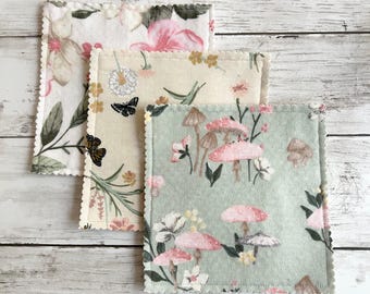 Op de afbeelding: Drie vierkante stoffen onderzetters met geschulpte randen. De bovenste onderzetter heeft een witte achtergrond met roze bloemen en groene bladeren. De middelste is beige met vlinders en paardenbloemen. De onderste is saliegroen met roze paddenstoelen.