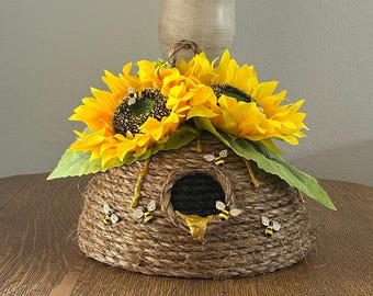 Op de afbeelding: Een decoratieve bijenkorf gemaakt van geweven touw, versierd met felgele zonnebloemen en groene bladeren. De korf heeft een donkere opening met een gouden honingdruppel en is versierd met kleine bijenversieringen.