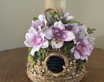 Op de afbeelding: Een decoratieve, geweven bijenkorf-vormige container met een ronde opening, versierd met faux paarse en witte bloemen en groene bladeren. De korf is versierd met kleine bijenbeeldjes. Bovenop zit een geweven handvat.